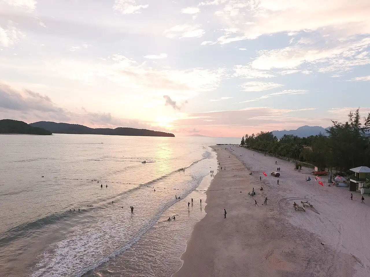 Aerial view of Cenang Beach at sunset, with people swimming in the ocean and walking on the sandy shore, framed by distant mountains