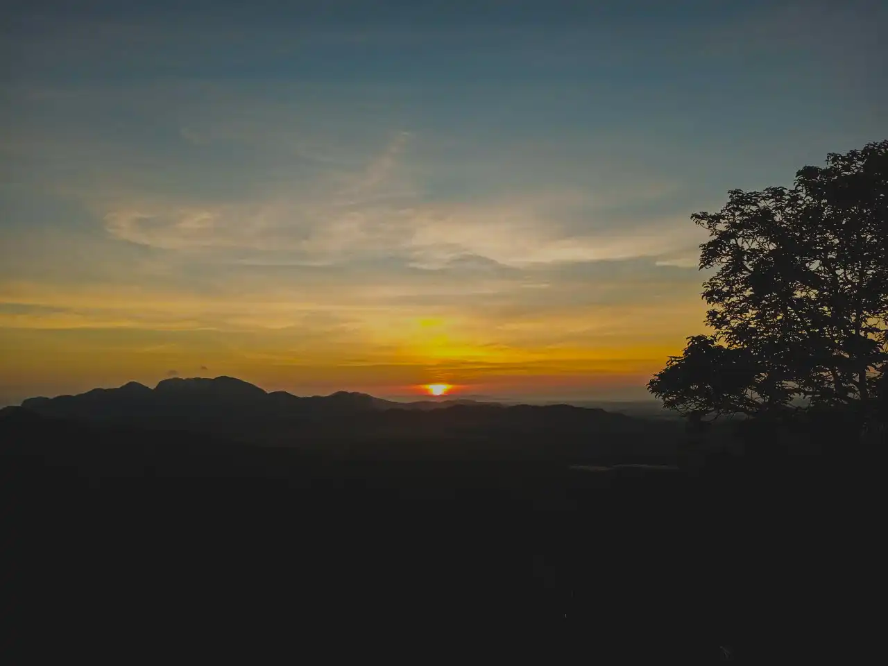 Vibrant sunset over the Wang Kelian mountains, with the sky glowing in orange and blue, silhouetting peaks and a large tree