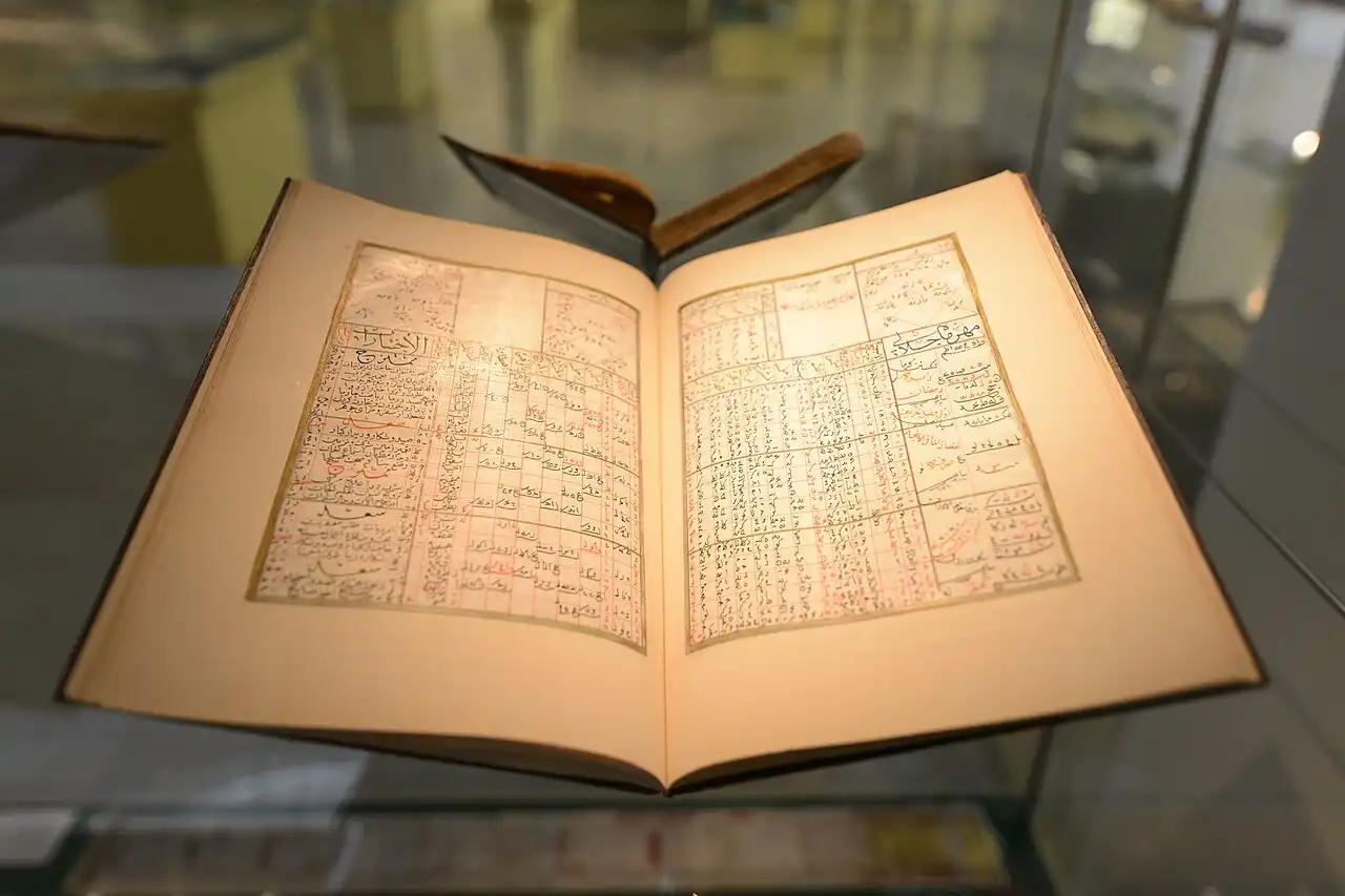 An open, antique book with detailed Arabic script and tables in black and red ink, showcased at Islamic Arts Museum Malaysia