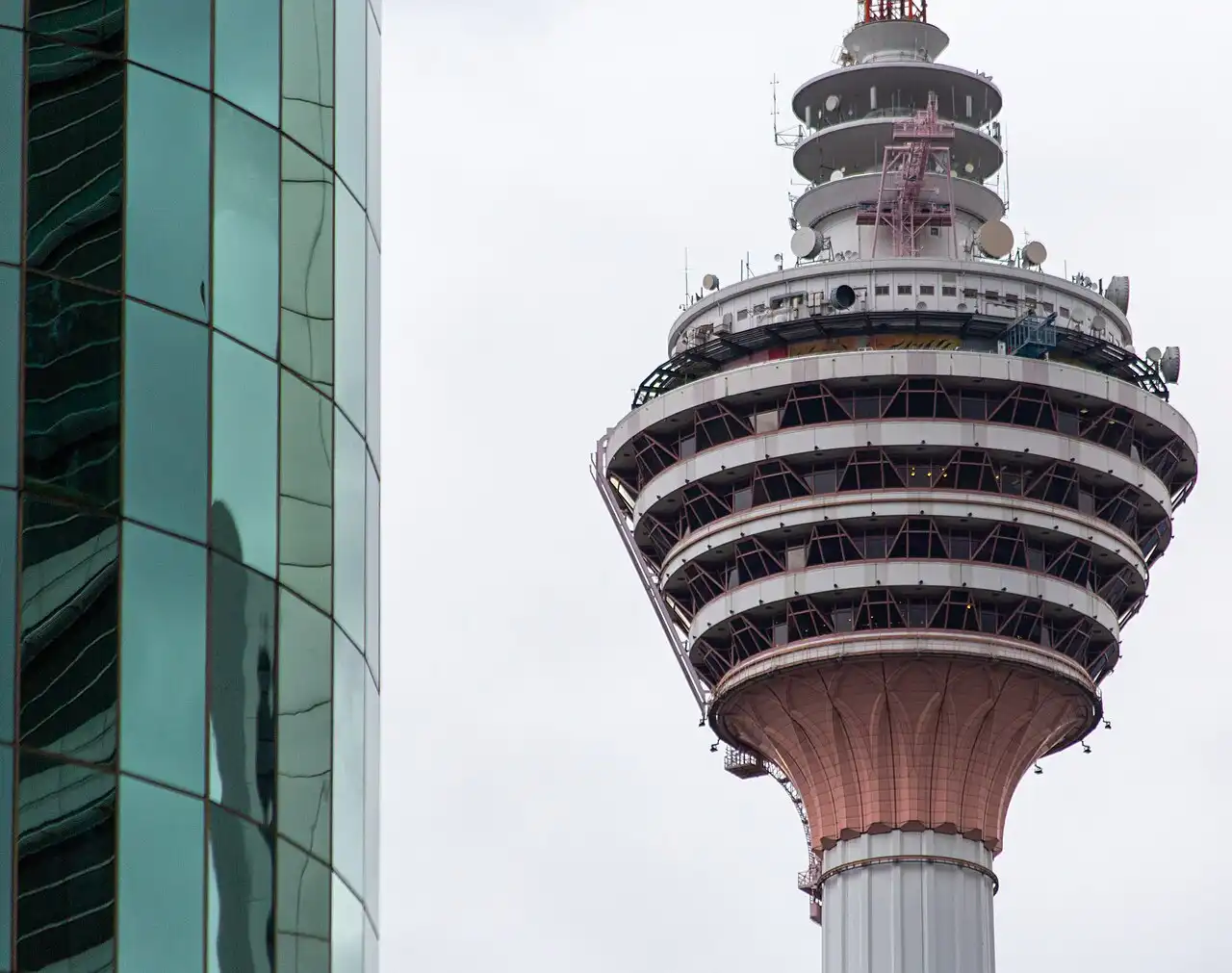 Close-up of the upper section of KL Tower with its distinctive observation deck and antennas, alongside a modern glass building