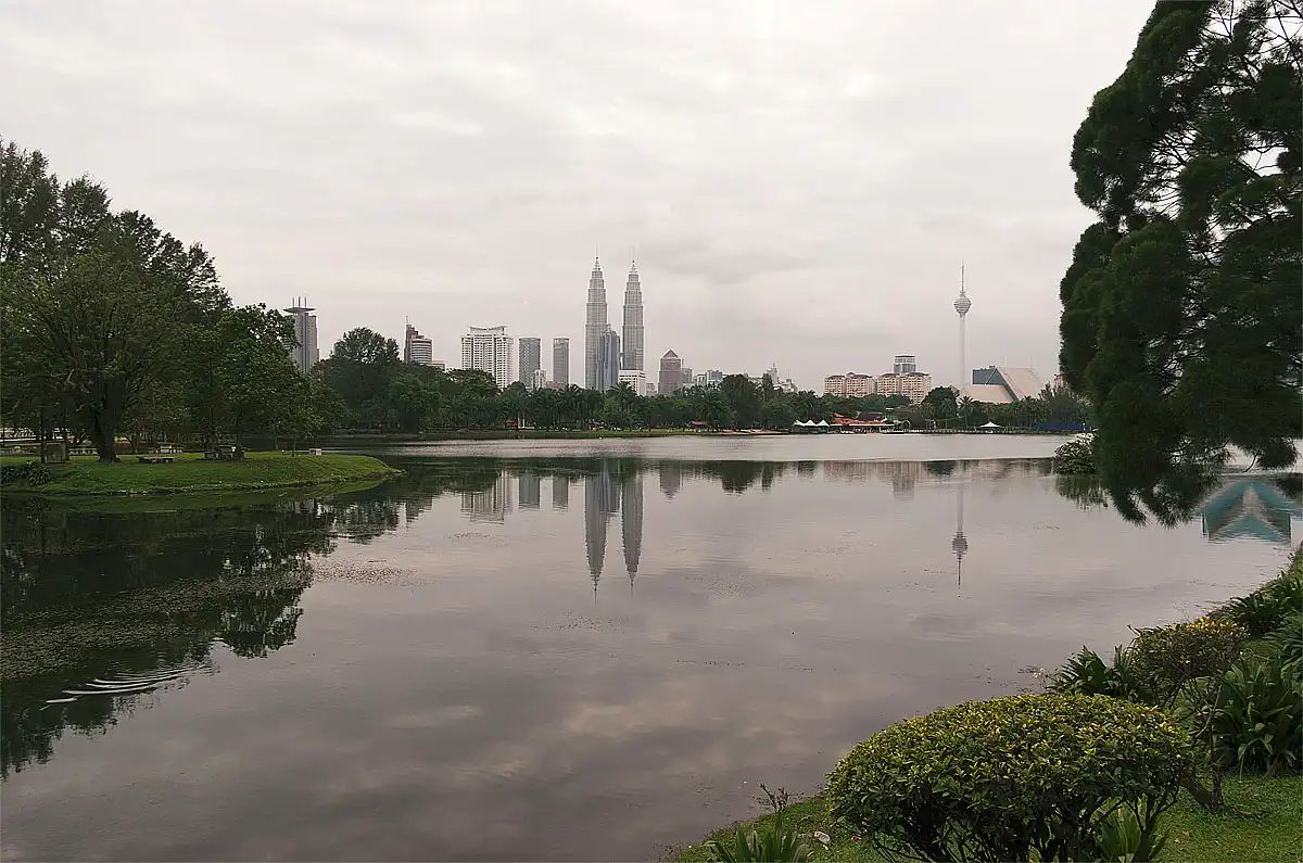 The Kuala Lumpur skyline, including the Petronas Twin Towers and KL Tower, reflected in the calm waters of Titiwangsa Lake Gardens