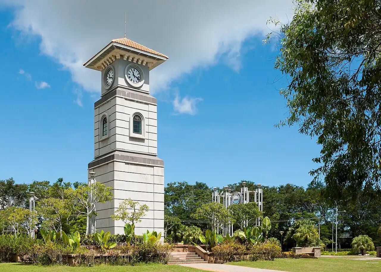 The iconic Labuan Clock Tower, a historic landmark, stands tall under a clear blue sky with lush greenery and a Labuan sign visible