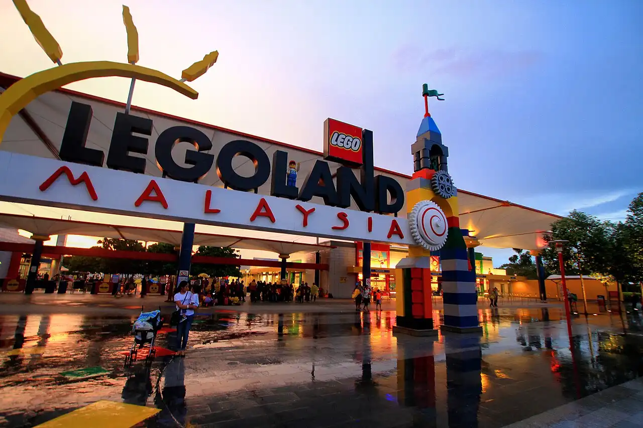 Legoland Malaysia park entrance at sunset, showing the large sign, brick tower, and visitors reflected on the wet ground