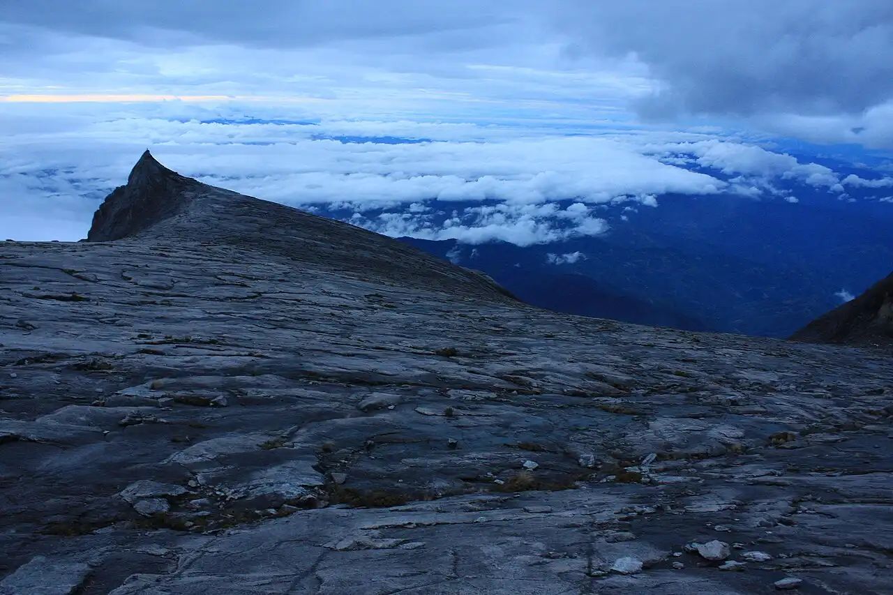Rocky summit of Mount Kinabalu rising above a sea of clouds at dawn, with distant peaks visible beneath a cloudy sky
