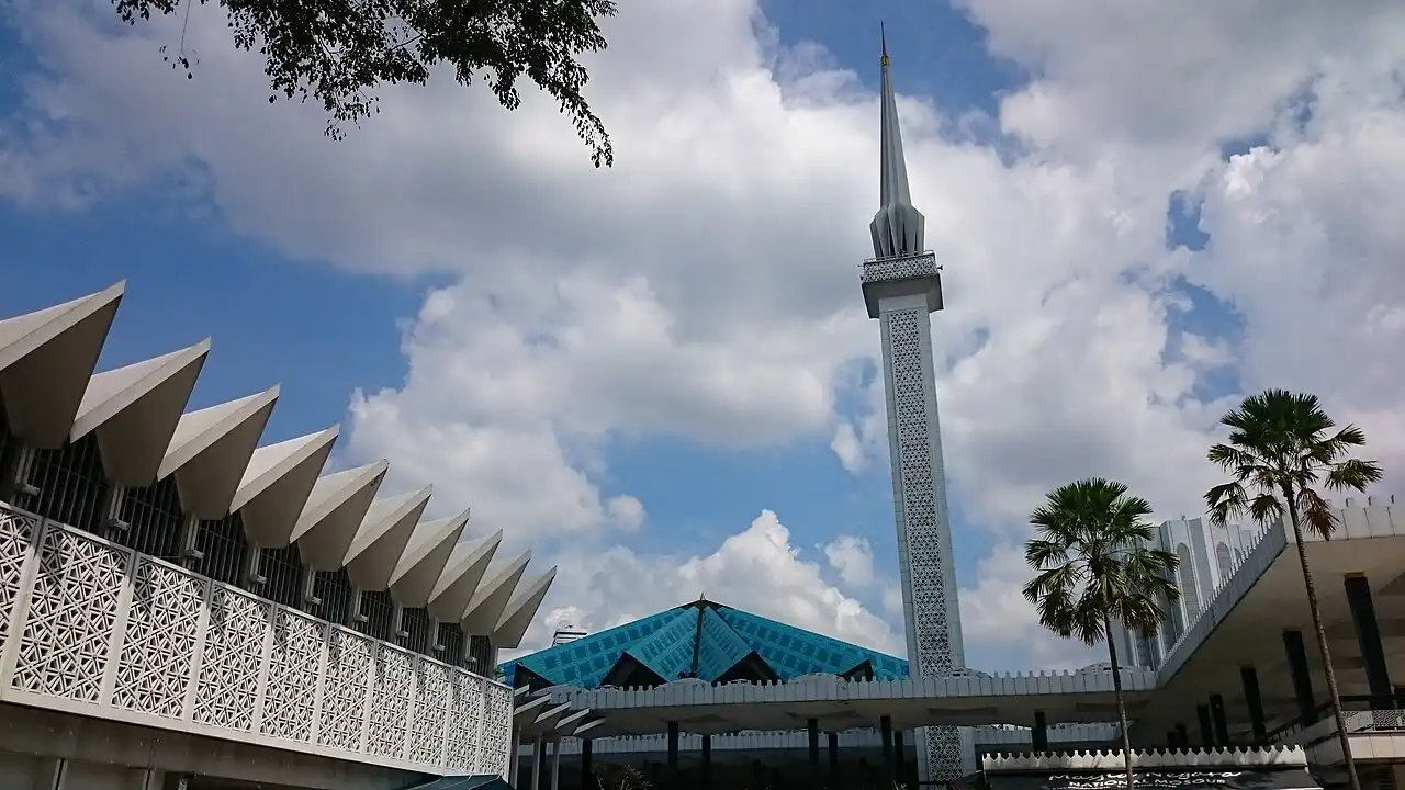 View of the National Mosque Kuala Lumpur's modern architecture, including its tall minaret, striking blue roof, and palm trees