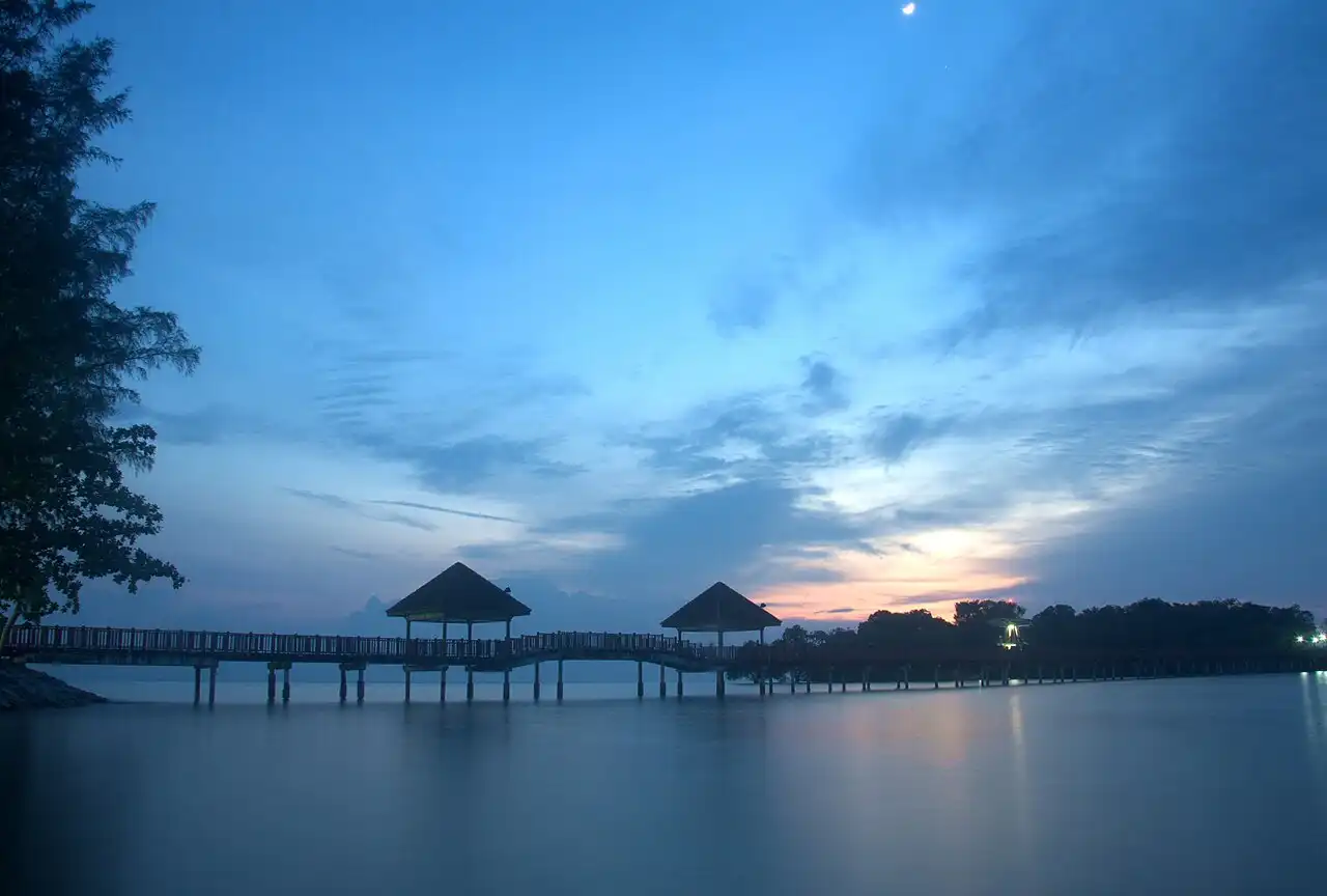 Tranquil twilight scene at Port Dickson featuring a wooden jetty with two gazebos over calm water and a crescent moon in the blue sky